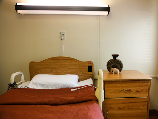 A simple hospital room featuring a wooden bed with white linens, a nightstand, and a decorative vase.