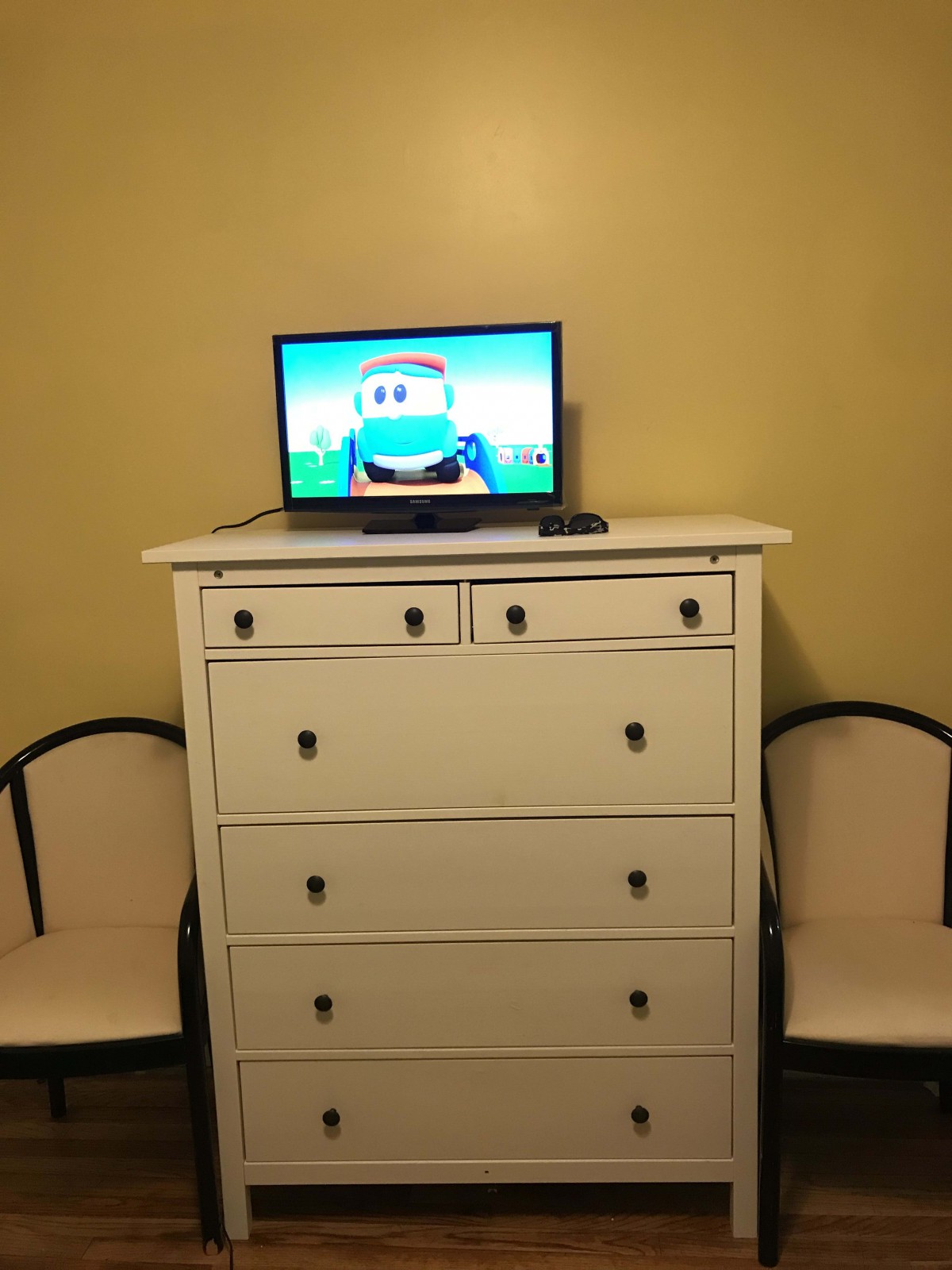 A small television on top of a white dresser with two chairs on either side, displaying a cartoon character.