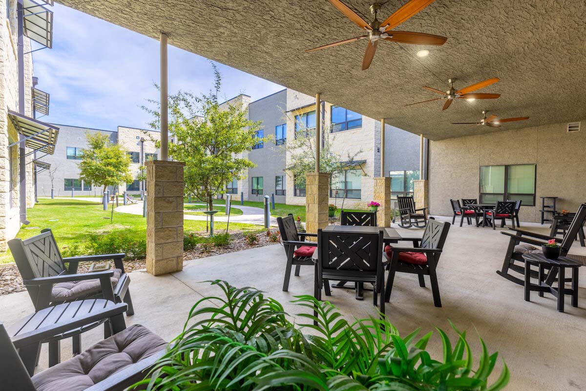 A modern outdoor patio with comfortable seating and ceiling fans, surrounded by greenery and contemporary buildings.