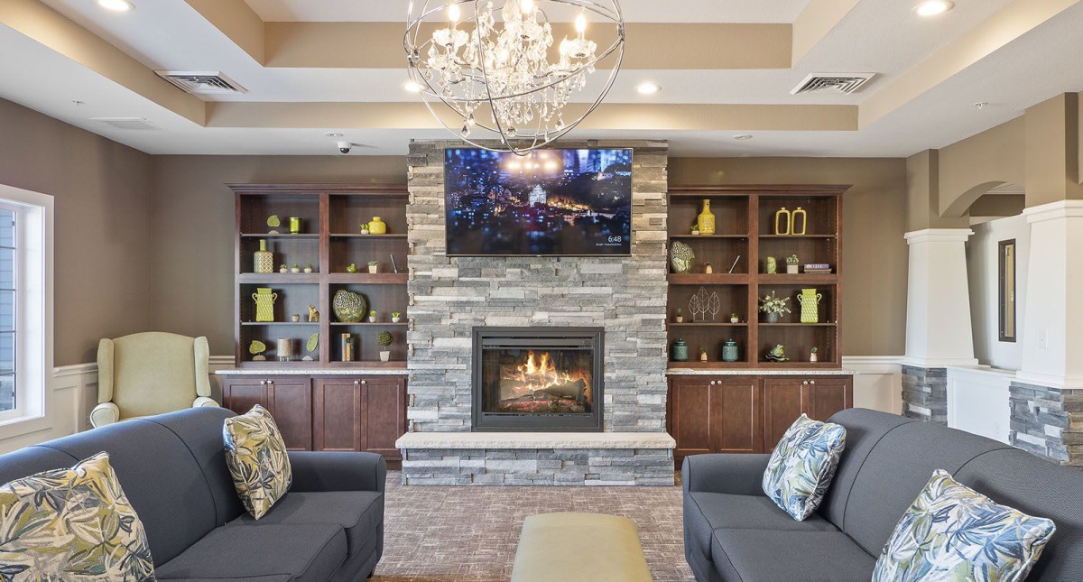 A cozy living room featuring a stone fireplace, a chandelier, and a TV mounted above the mantel. The room is furnished with comfortable couches and decorative shelves.