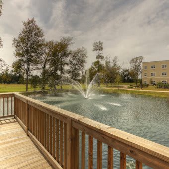 A peaceful view of a pond with a fountain, surrounded by trees and overlooked by a wooden bridge.