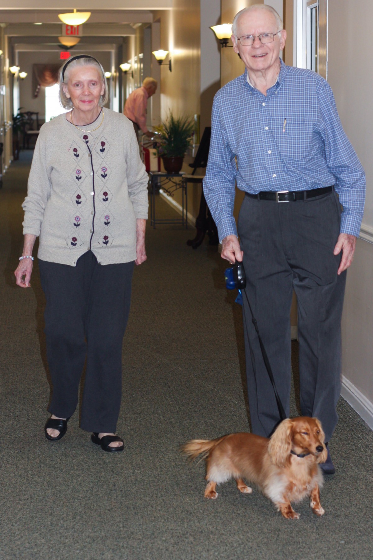An elderly couple walking their dog indoors.