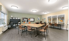 A modern classroom with tables and chairs arranged for group activities, featuring decorative elements and a bulletin board.