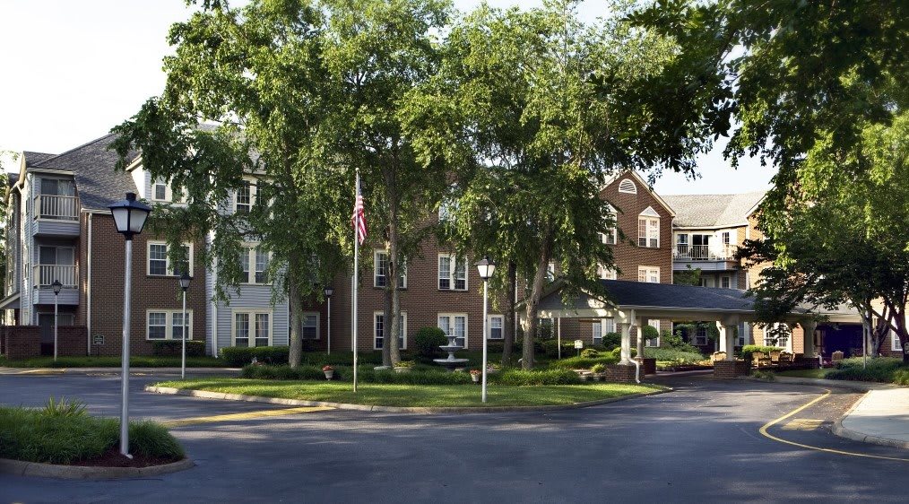 A large brick building surrounded by trees, featuring a driveway with a flagpole at the entrance.