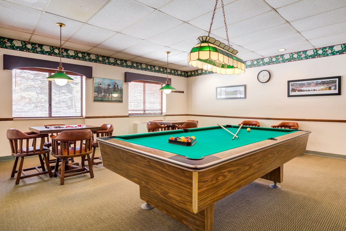 A cozy billiards room with a classic pool table, surrounded by wooden chairs and tables, under warm lighting.