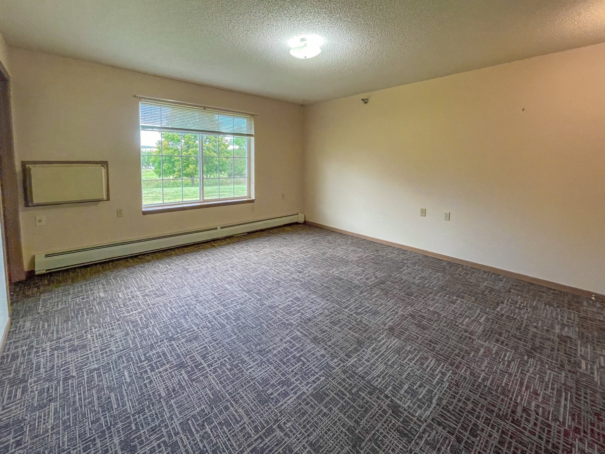 An empty, carpeted room with a large window and plain walls.