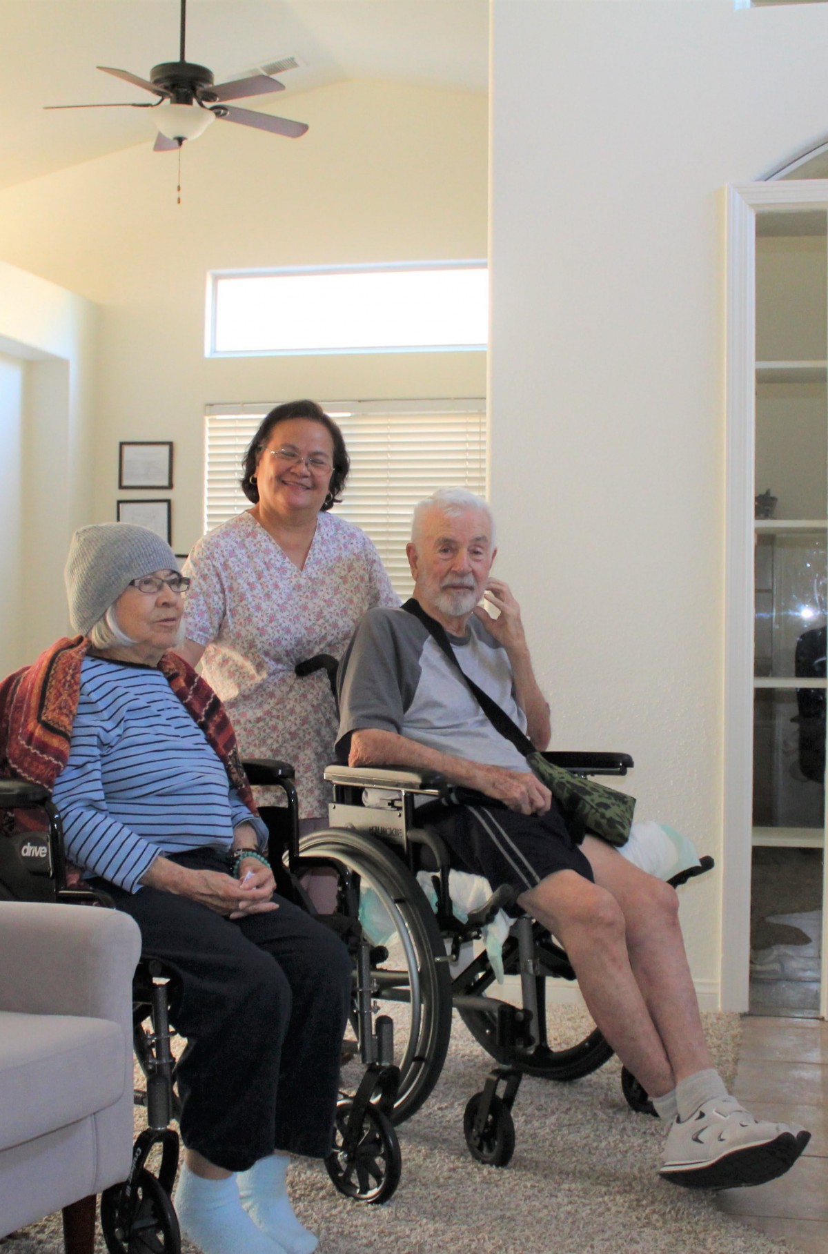 An elderly couple in wheelchairs, accompanied by a caregiver, in a welcoming indoor environment.