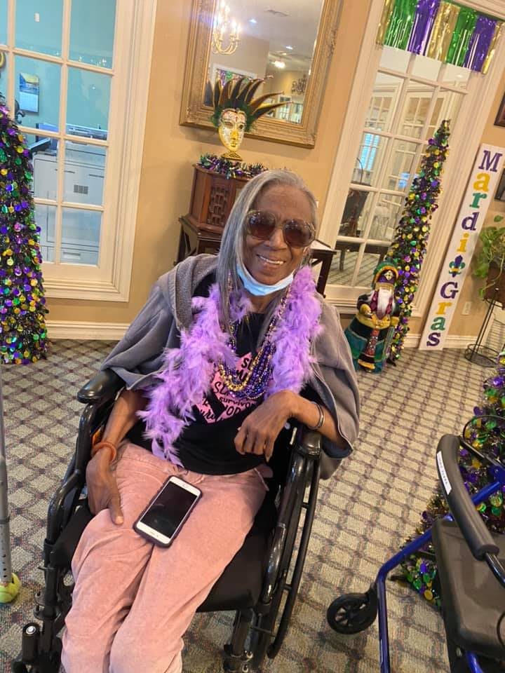 An elderly person wearing a scarf, sunglasses, and a purple boa is seated in a wheelchair indoors.