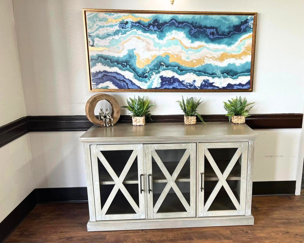 A stylish console table with glass front cabinets, decorated with small plants and a decorative piece, is situated beneath a wavy abstract artwork.