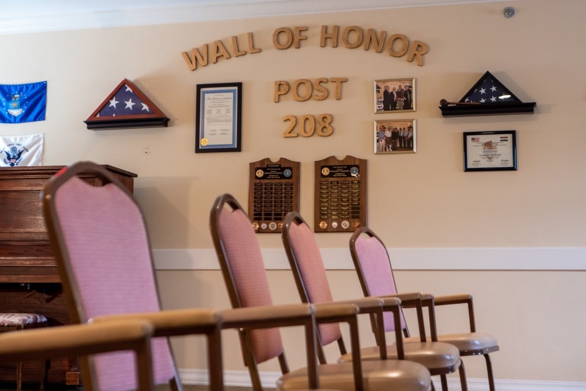 A Wall of Honor at Post 208 featuring plaques, memorial flags, and chairs arranged in a room.