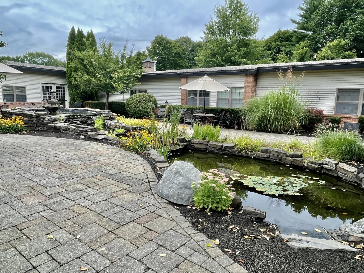 A serene garden featuring a pond surrounded by lush greenery and a brick patio.