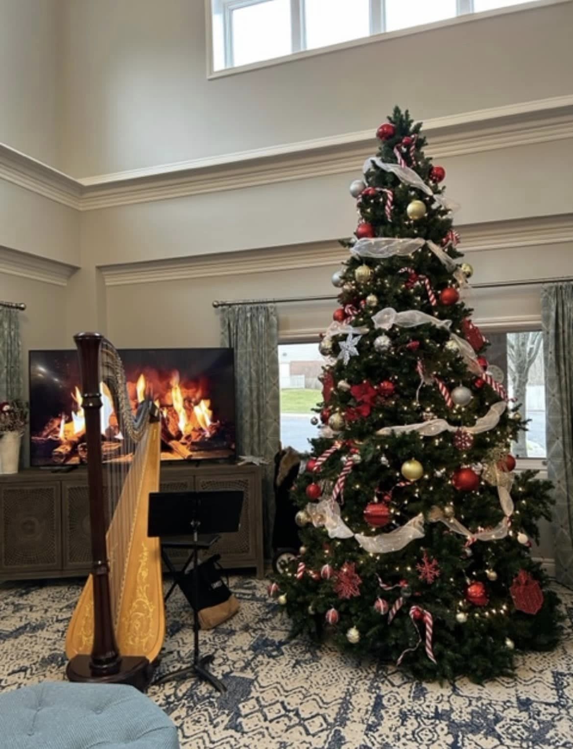 A beautifully decorated Christmas tree stands next to a harp, with a cozy fireplace on the TV creating a festive holiday atmosphere.