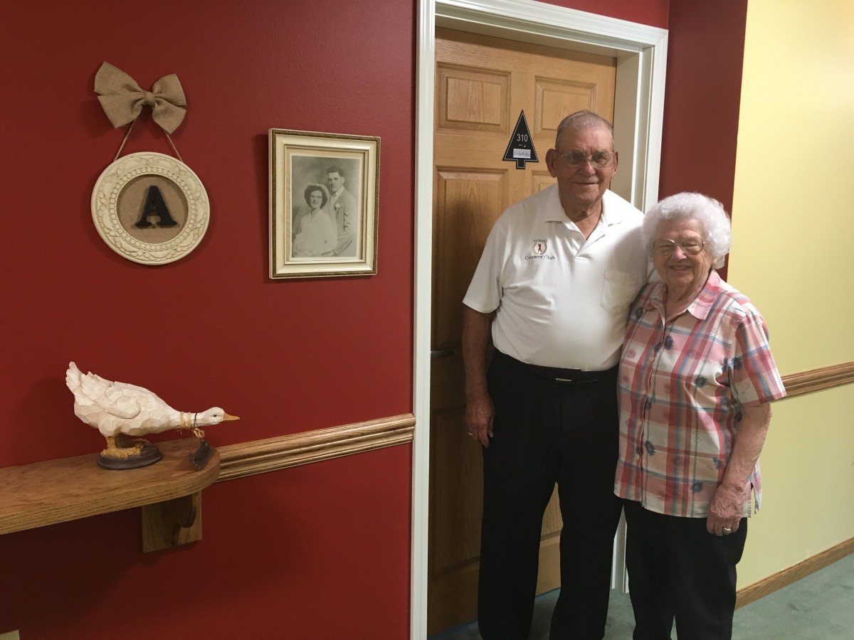 An elderly couple stands together in a warmly decorated room with red walls.