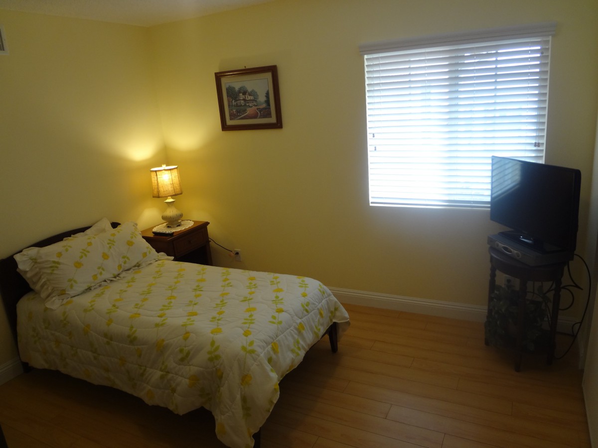 A cozy bedroom featuring a single bed, a bedside lamp, a window with blinds, and a television on a stand.