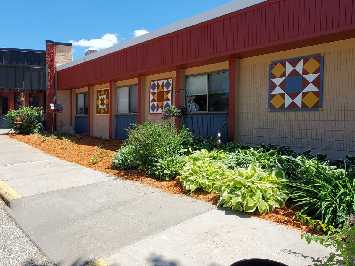 A sunny view of a building exterior decorated with quilt patterns and surrounded by landscaped greenery.