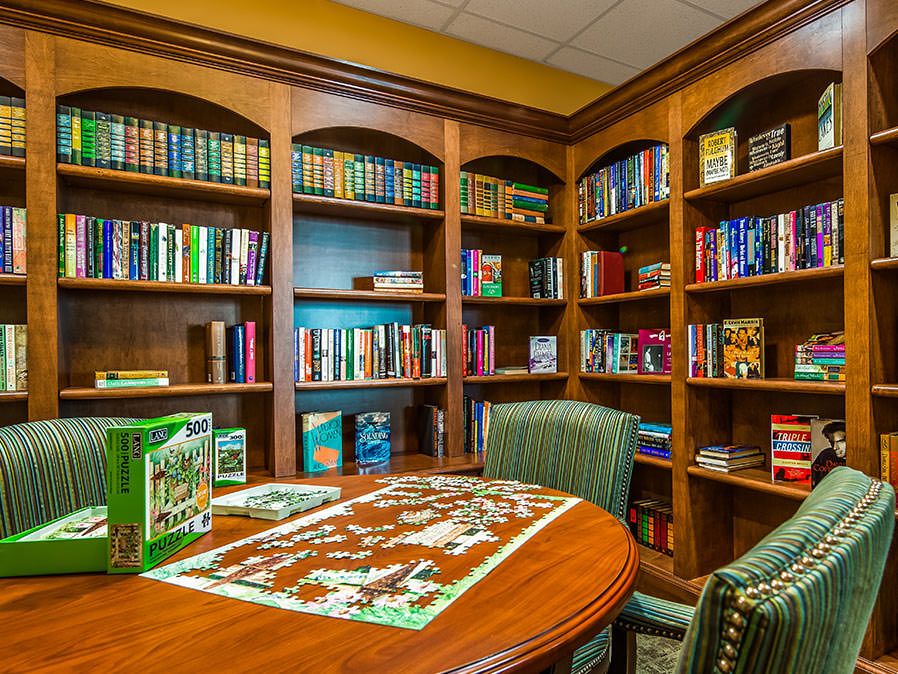 A cozy library corner with wooden bookshelves filled with books, a round table and chairs, and a jigsaw puzzle in progress.