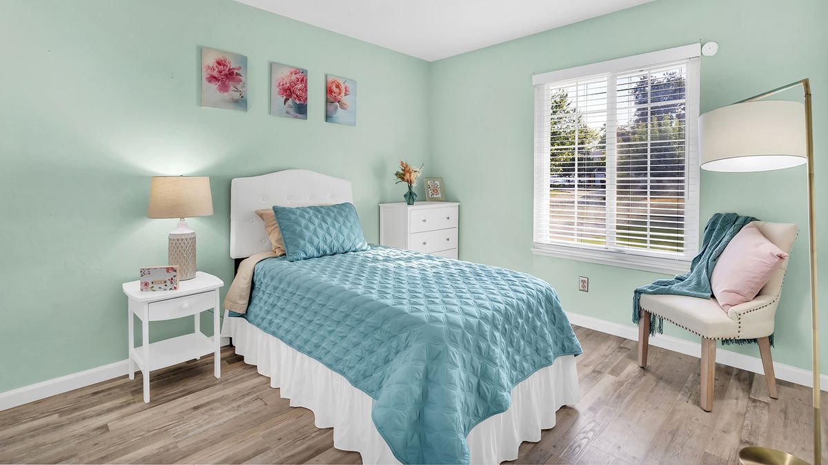 A cozy bedroom with a single bed covered in blue bedding, complemented by floral wall decor and a view of the outside through the window.