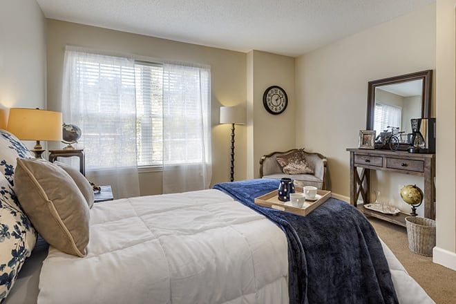 A cozy bedroom with elegant decor featuring a neatly made bed, a plush armchair, and tasteful furnishings including a lamp and mirror.