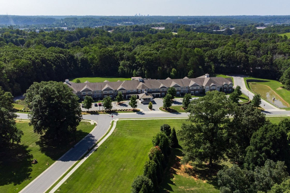 Aerial view of a building surrounded by greenery and trees, with a visible roadway and parking lot.