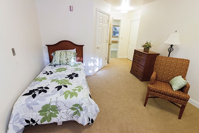 A cozy, well-furnished bedroom featuring a single bed with patterned bedding, a wooden dresser, and an armchair.