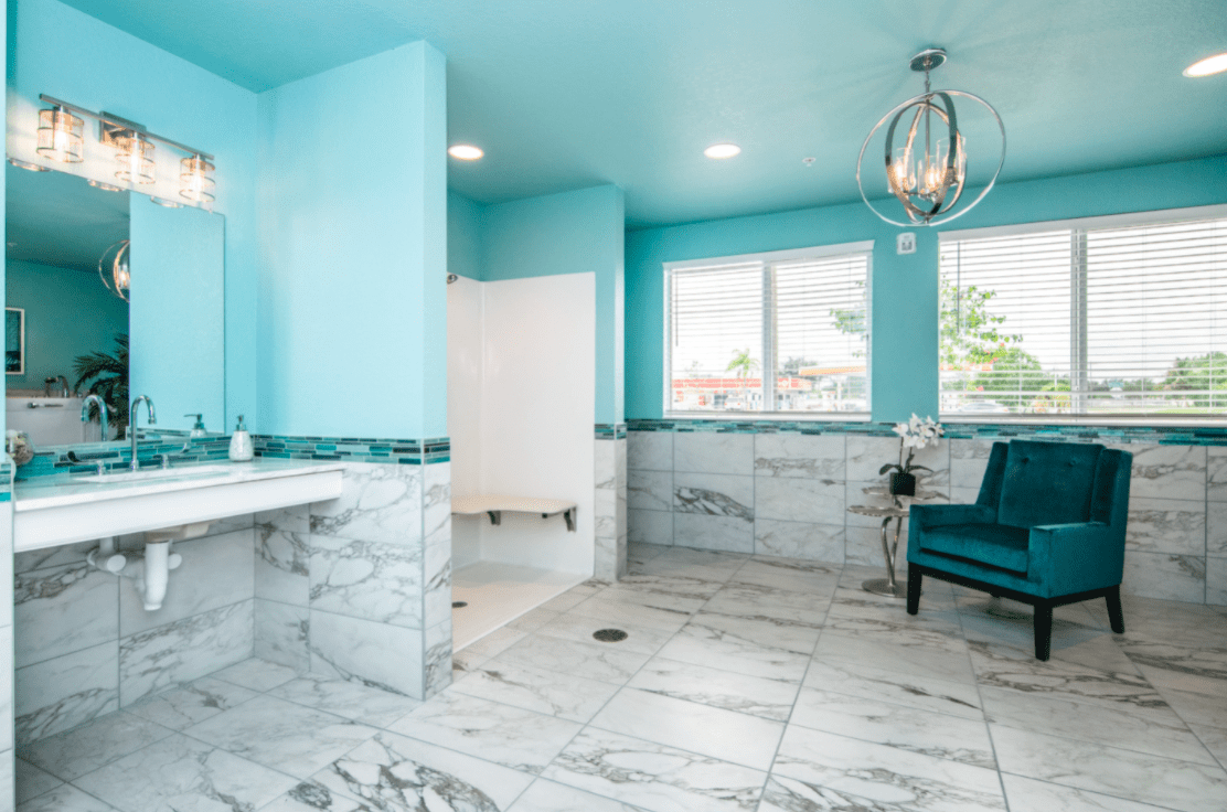 A modern bathroom with marble flooring and turquoise walls, featuring a luxurious armchair and chandelier.