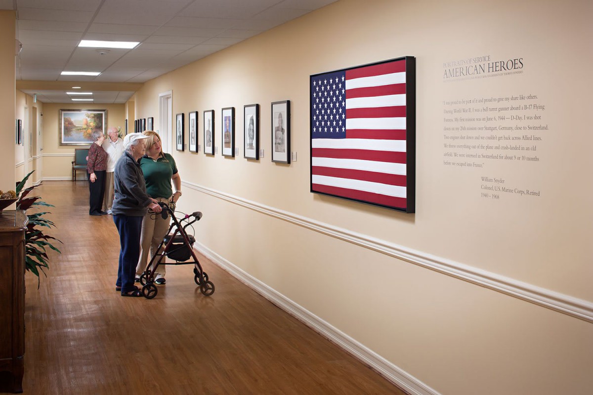 An elderly couple admires a framed American flag and artwork displayed in a hallway.
