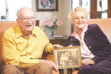 An elderly couple smiling while holding an old photograph in their living room.