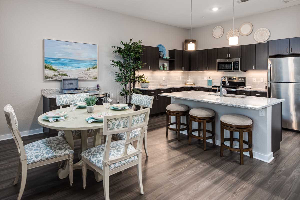 A modern kitchen and dining area featuring a sleek design with dark cabinetry, an island with bar stools, and a round dining table set. The space is complemented by an indoor plant and coastal artwork.