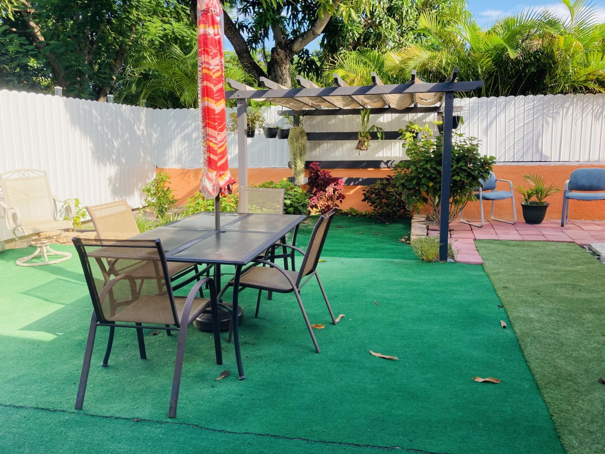 A cozy backyard patio featuring a table with chairs under a colorful umbrella, adjacent to a pergola surrounded by lush greenery.