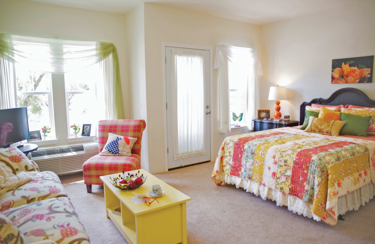 A bright and cozy bedroom with colorful decor, featuring a bed with a patchwork quilt, an armchair, and natural light coming in through the windows.