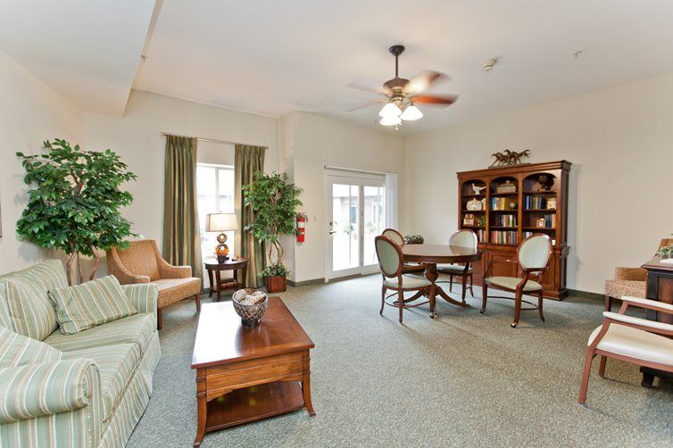 A cozy living room with traditional furniture, featuring a table and chairs, a bookshelf, indoor plants, and large windows with curtains.