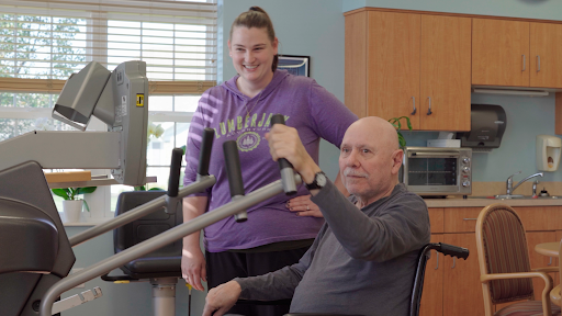 A senior man engages in a strength training exercise with the assistance of a trainer in a rehabilitation setting.