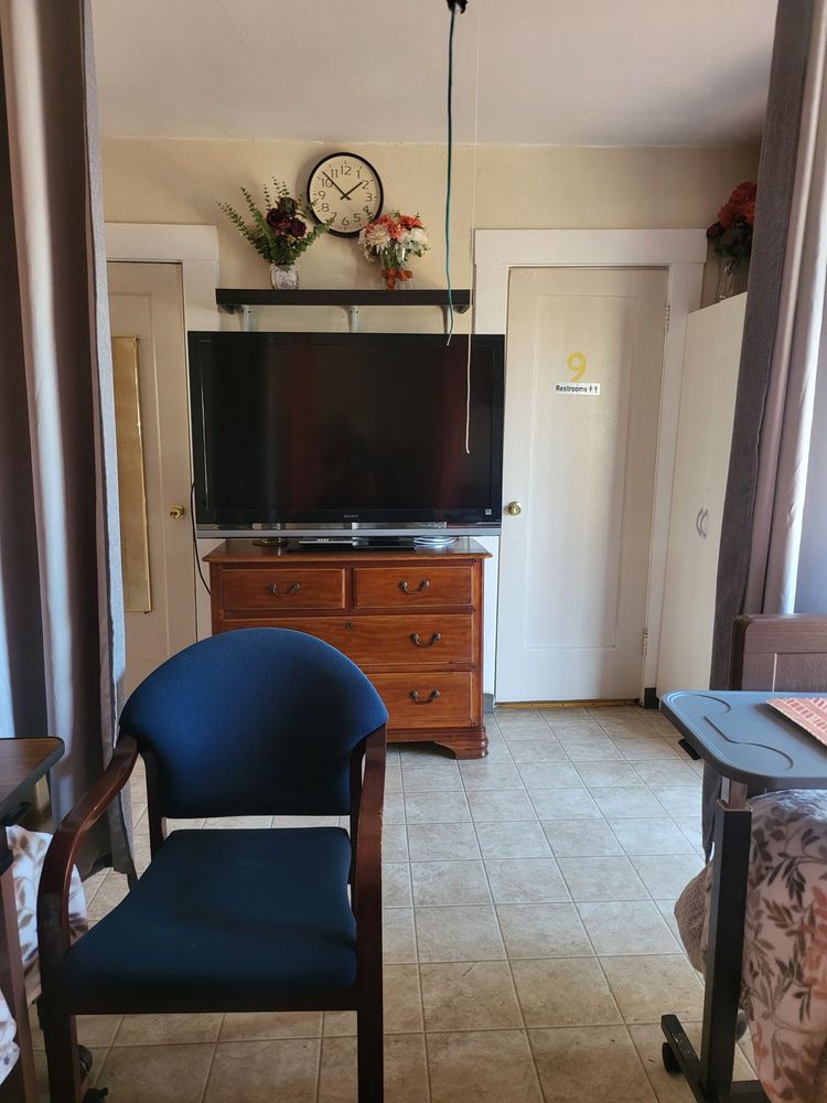 A cozy living room with a television on top of a wooden dresser, a navy blue chair in the foreground, and decorative elements including a clock and flowers.