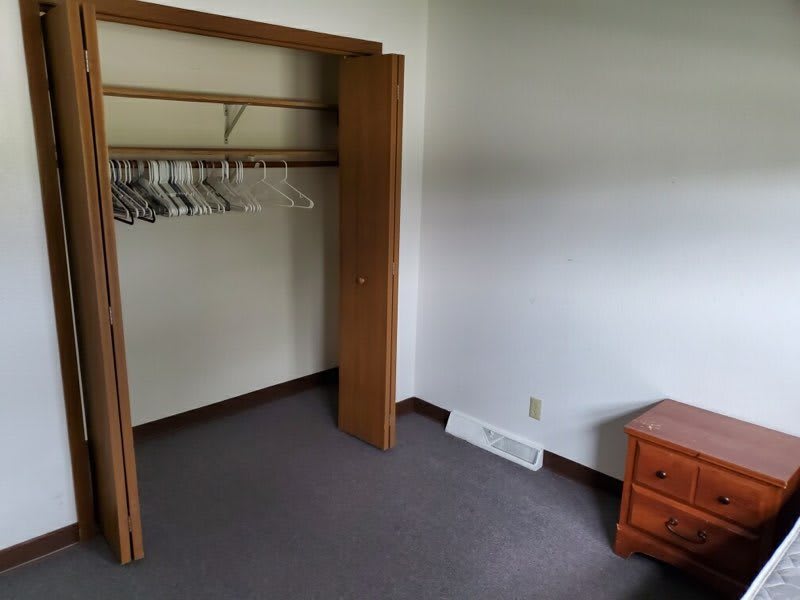 An empty room with an open closet containing hangers and a wooden nightstand.