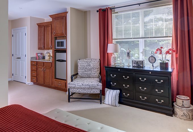 A cozy living room featuring a dresser, patterned chair, and a large window with red curtains.
