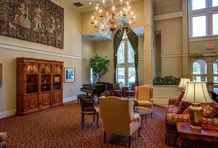 A spacious and elegant living room featuring classic furniture, a grand piano, a large chandelier, and a cozy seating area with bookshelves.