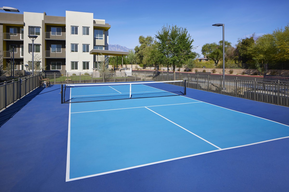 An outdoor blue tennis court surrounded by a fence near a residential building on a sunny day.