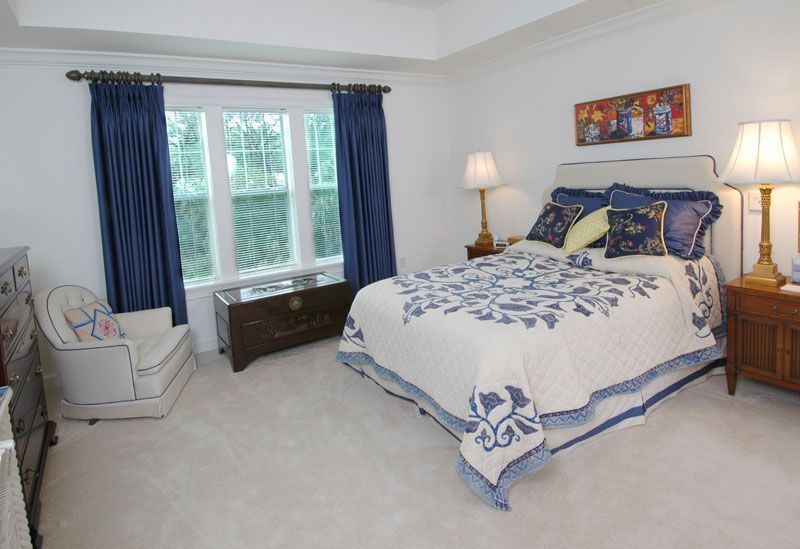 A cozy bedroom with blue and white decor, featuring a neatly made bed, a window with blue curtains, and a bedside lamp.
