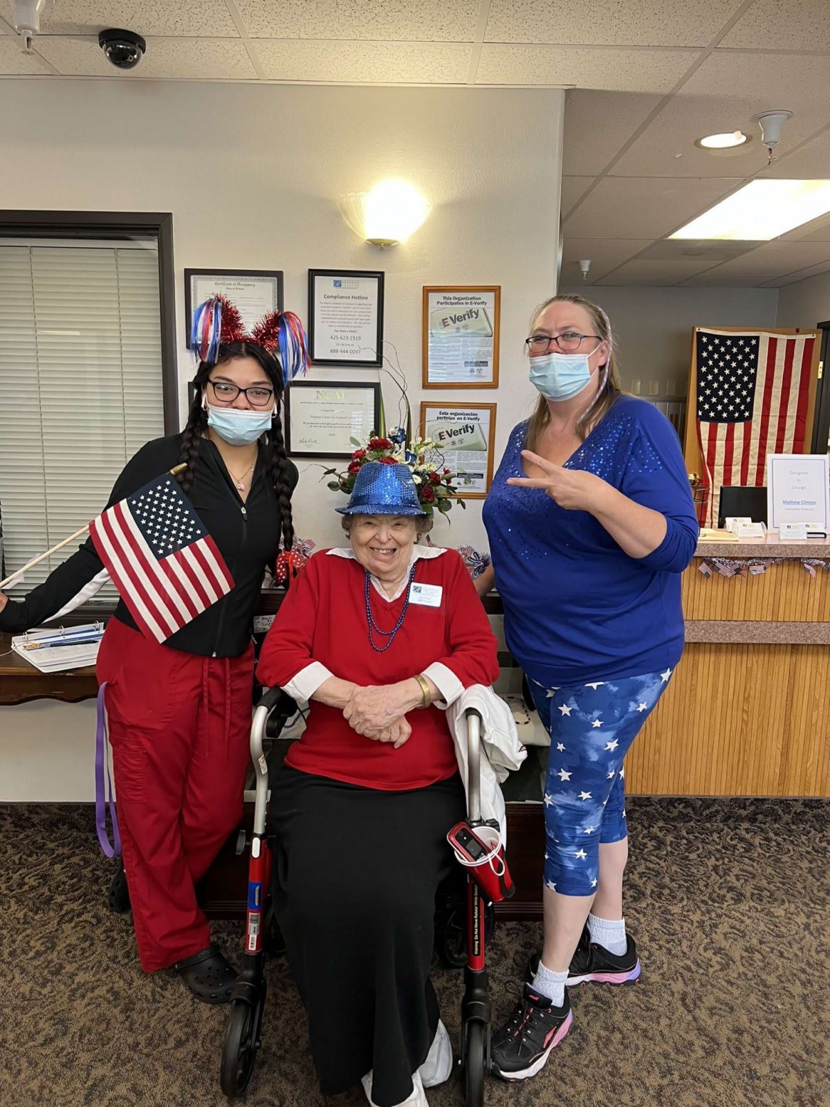 A group of three people celebrating a patriotic event indoors, with one holding an American flag. They are all wearing masks and festive attire.