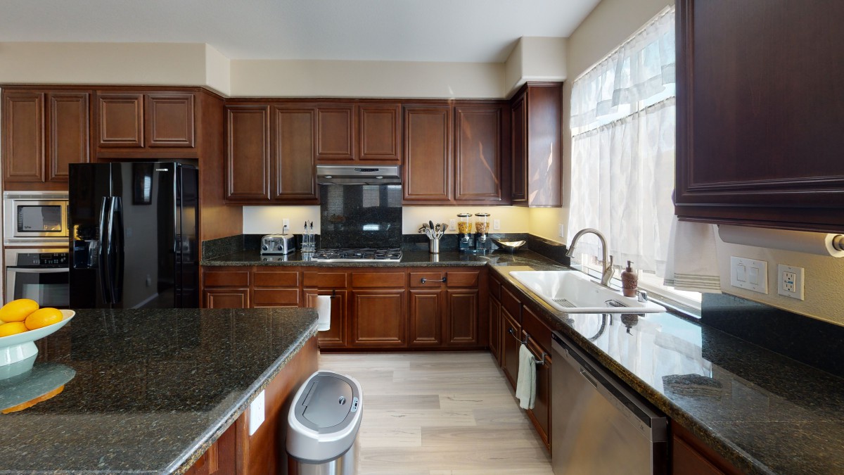 A modern kitchen with wooden cabinets and black countertops, featuring a sink, stove, oven, and ample natural light.