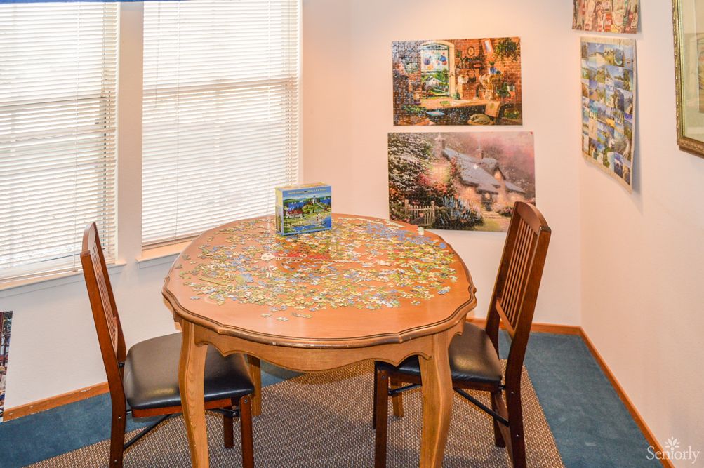 A cozy room with a wooden table featuring an incomplete jigsaw puzzle, surrounded by chairs and decorated with artwork on the walls.