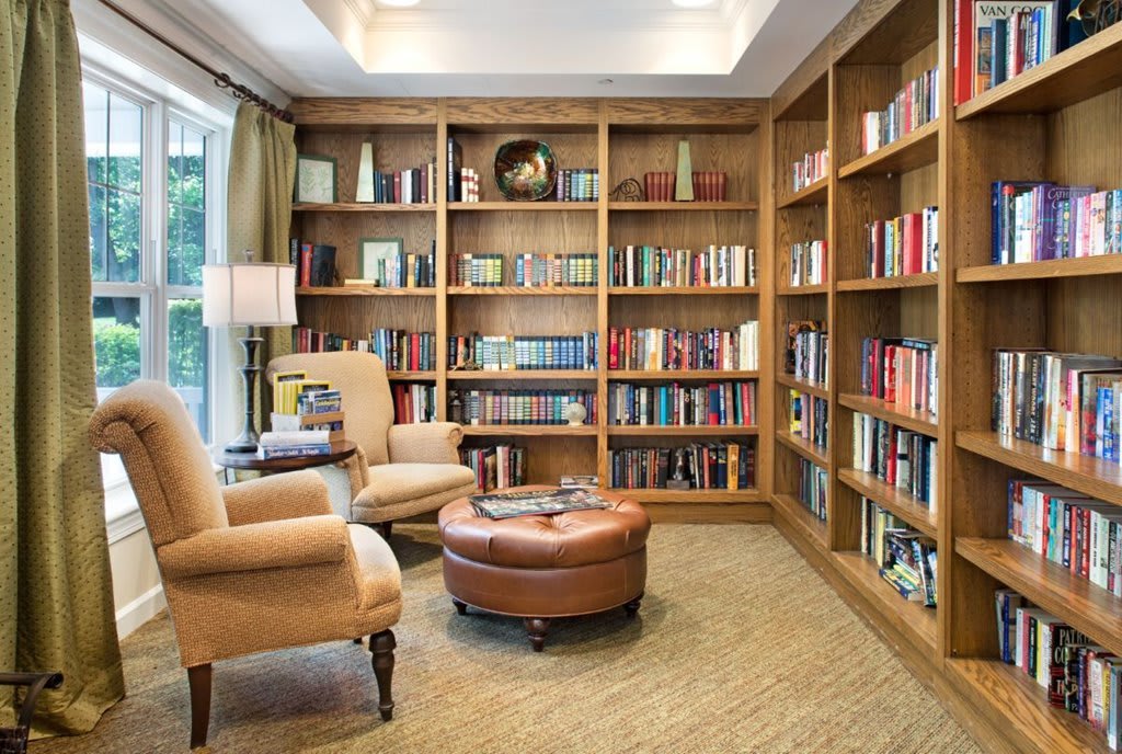 A cozy home library with wooden bookshelves filled with books, comfortable chairs, and a round leather ottoman.
