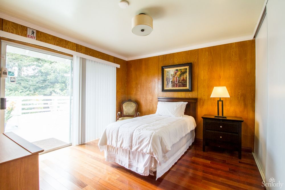 A cozy bedroom with wood paneling, featuring a single bed, nightstand with lamp, and sliding door leading outside.