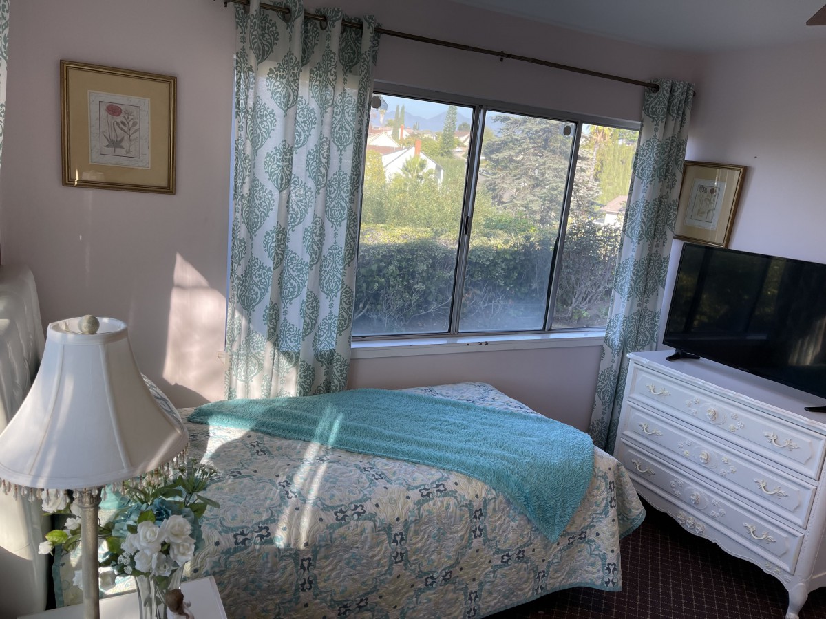 A cozy bedroom with a bed near a large window, featuring patterned curtains and a TV on the dresser.