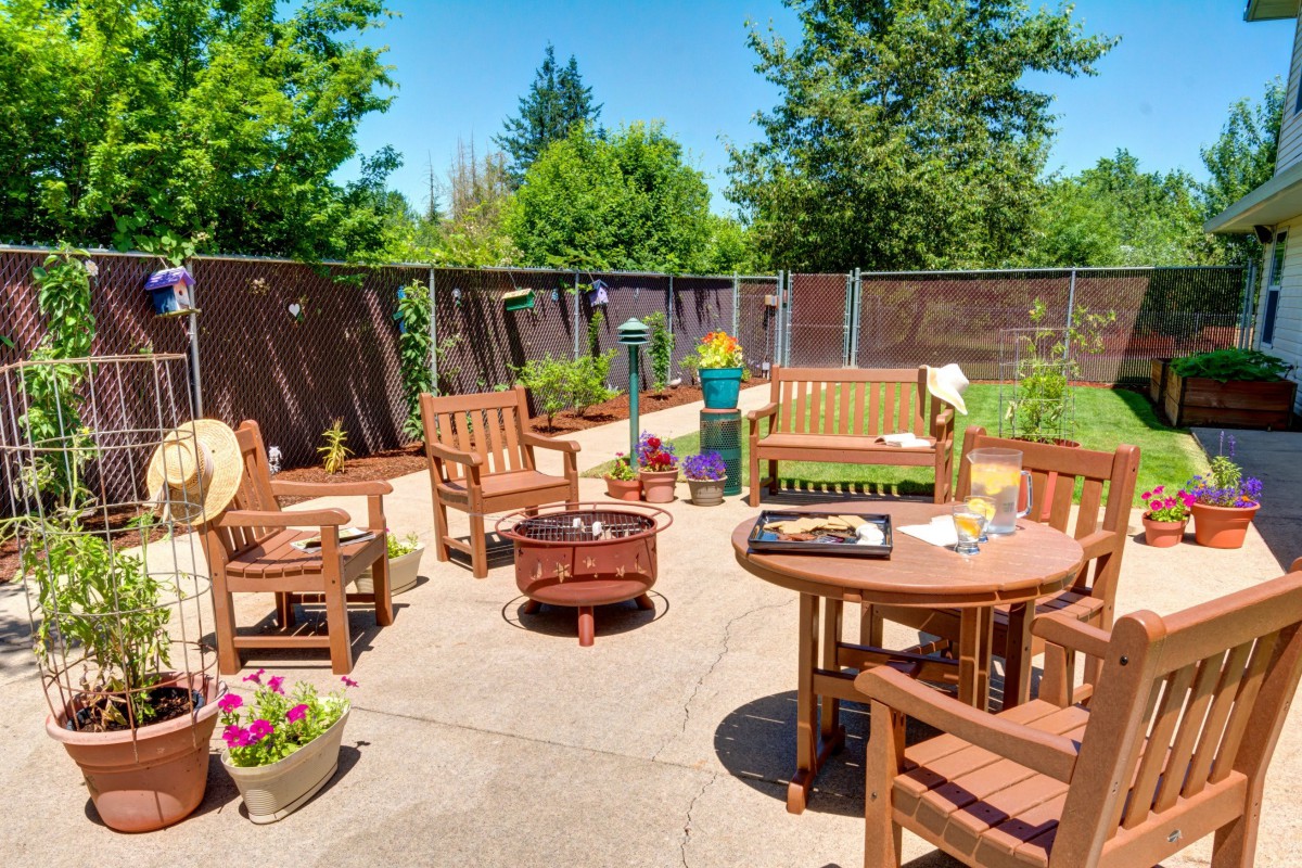 A sunny patio area with wooden furniture and a fire pit, surrounded by lush greenery.