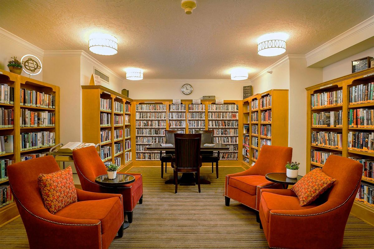 A cozy library reading room with bookshelves, orange chairs, and a central table.