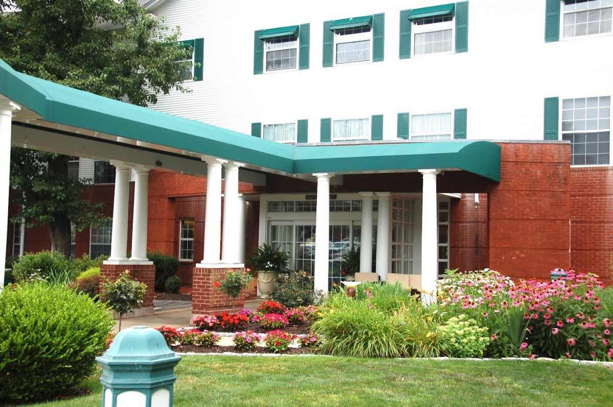 An exterior view of a building entrance with green canopies, columns, and landscaped garden.