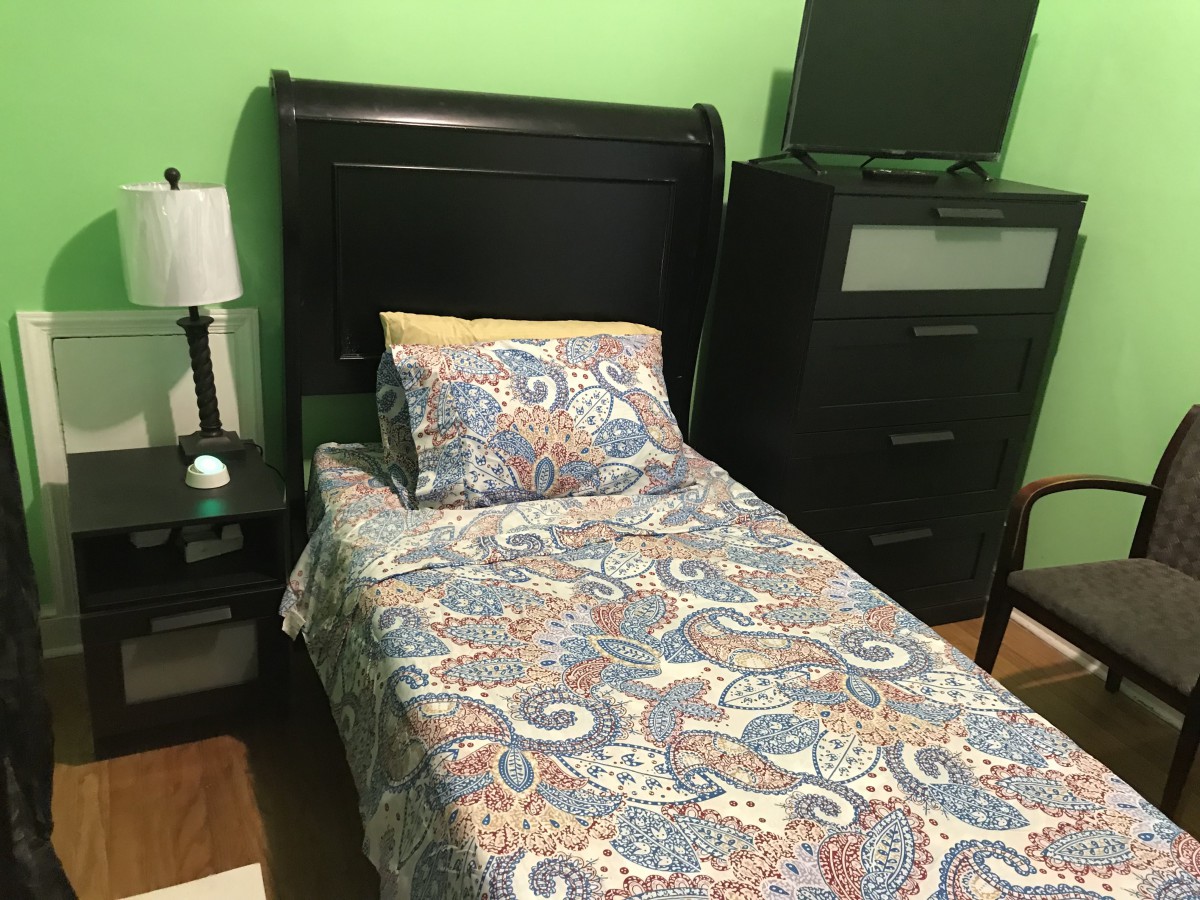 A cozy bedroom with a single bed featuring colorful bedding and a dark wooden headboard, next to a nightstand with a lamp and a tall dresser against a green wall.