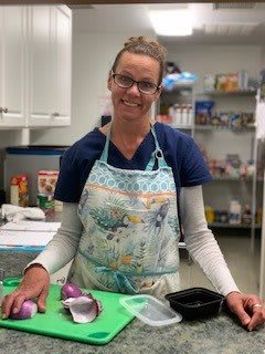 A woman wearing an apron and glasses smiles while standing in a kitchen setting.