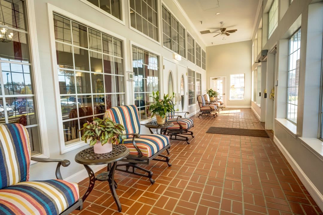 A bright indoor lounge area with striped chairs, small tables, and potted plants lined along large windows.
